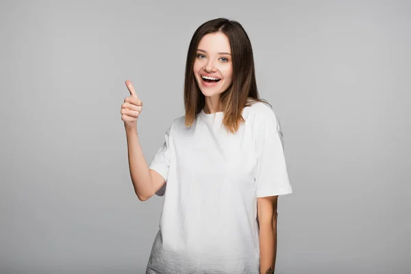 Happy brunette woman showing thumb up while looking at camera isolated on grey — Stock Photo