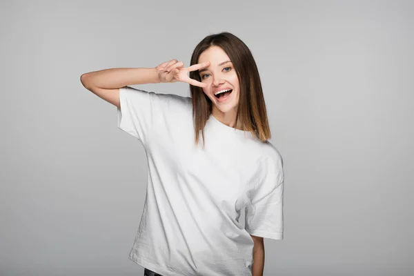 Eccitato bruna donna mostrando segno di vittoria vicino al volto isolato sul grigio — Foto stock