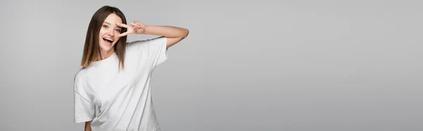 Fröhliche Frau in weißem T-Shirt mit Siegeszeichen in Gesichtsnähe isoliert auf grauem Banner — Stockfoto