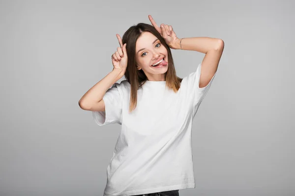 Mujer alegre y difícil sobresaliendo lengua y mostrando los cuernos gesto aislado en gris - foto de stock