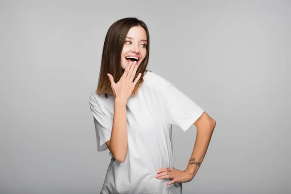 Laughing woman covering mouth with hand while standing with hand on hip isolated on grey — Stock Photo