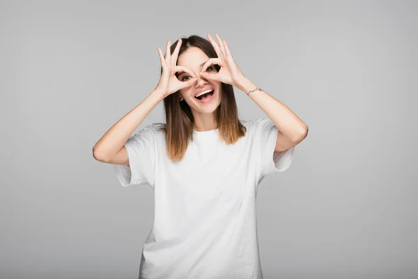 Mujer alegre en camiseta blanca que muestra signos de bien cerca de los ojos aislados en gris - foto de stock