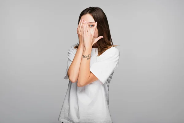Scared woman obscuring face with hands and looking at camera isolated on grey — Stock Photo
