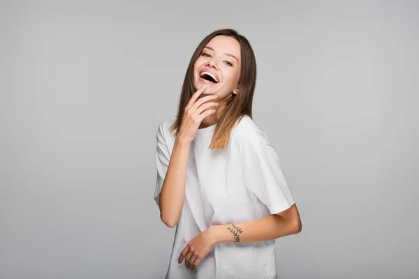 Збуджена жінка в білій футболці сміється, дивлячись на камеру ізольовано на сірому — стокове фото