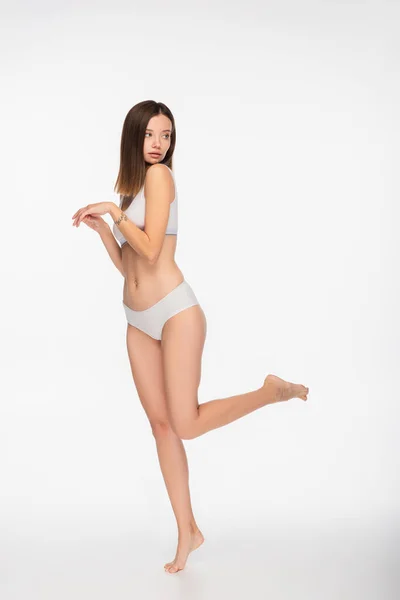 Vista completa de la mujer joven en lencería mirando hacia atrás mientras posa sobre una pierna sobre fondo blanco - foto de stock