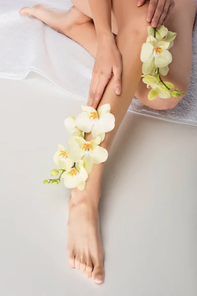 Abgeschnittene Ansicht einer barfüßigen Frau mit glatter Haut, die auf einem weißen Handtuch mit Orchideen sitzt — Stockfoto