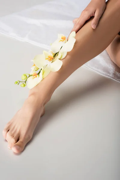 Cropped view of barefoot woman with orchid branch on leg sitting on white towel — Stock Photo