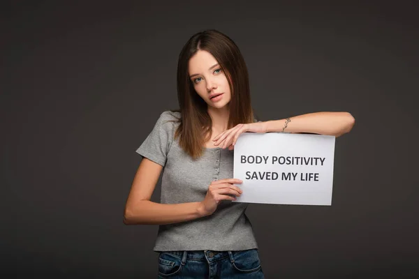 Mulher bonita olhando para a câmera perto do cartão com positividade do corpo salvou a minha vida lettering isolado em cinza — Fotografia de Stock