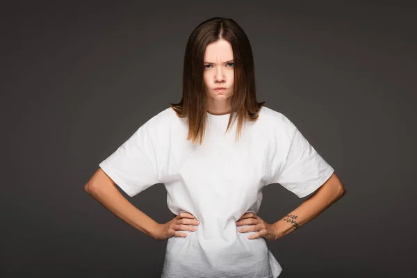 Angry woman puffing cheeks while standing akimbo isolated on dark grey — Stock Photo