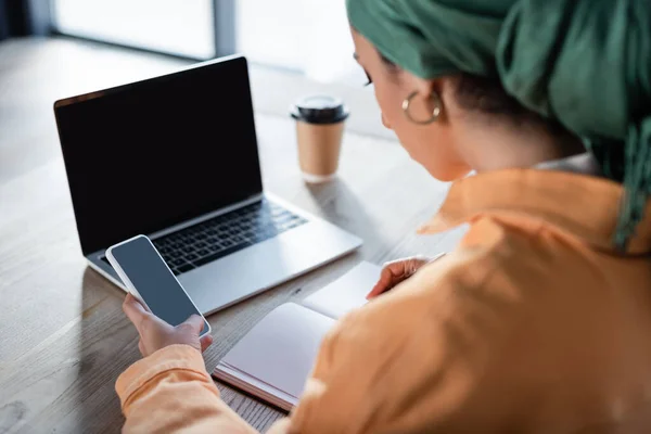 Mujer musulmana borrosa sosteniendo teléfono inteligente cerca de portátil y portátil con pantalla en blanco - foto de stock