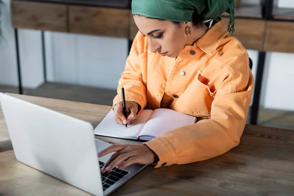 Mulher muçulmana escrevendo em notebook em branco ao usar laptop em casa — Fotografia de Stock