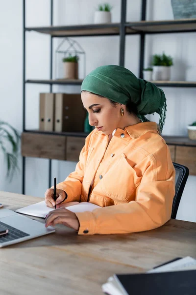 Muslimische Frau in orangefarbener Jacke und Kopftuch schreibt in Notizbuch, während sie zu Hause studiert — Stockfoto