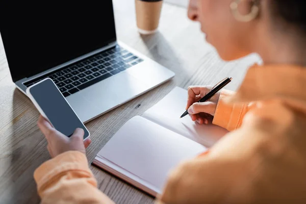 Vista recortada de la mujer borrosa con la escritura de teléfonos inteligentes en un cuaderno vacío cerca de la computadora portátil con pantalla en blanco - foto de stock