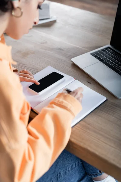 Blurred arabian woman using smartphone and writing in notebook while learning at home — Stock Photo