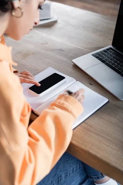 Mujer árabe borrosa usando un teléfono inteligente y escribiendo en un cuaderno mientras aprende en casa - foto de stock