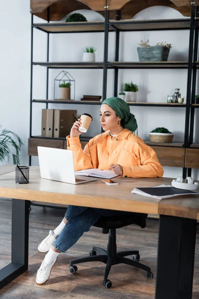 Mujer musulmana sosteniendo taza de papel mientras está sentado cerca de la computadora portátil en casa - foto de stock