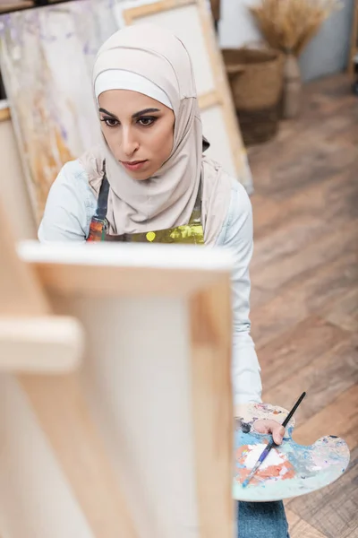 Hochwinkelaufnahme einer muslimischen Frau in der Nähe einer verschwommenen Staffelei zu Hause — Stockfoto