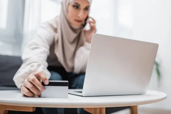 Selektiver Fokus der Kreditkarte in der Hand einer muslimischen Frau, die in der Nähe ihres Laptops mit dem Handy spricht — Stockfoto