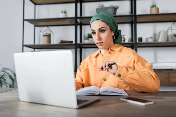 Mulher muçulmana em headkerchief e jaqueta laranja sentado com caneta perto de caderno borrado — Fotografia de Stock
