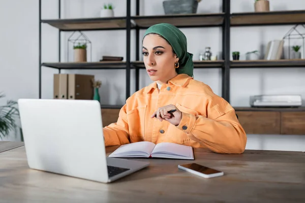 Mujer árabe en el pañuelo mirando portátil cerca de cuaderno vacío en el escritorio - foto de stock