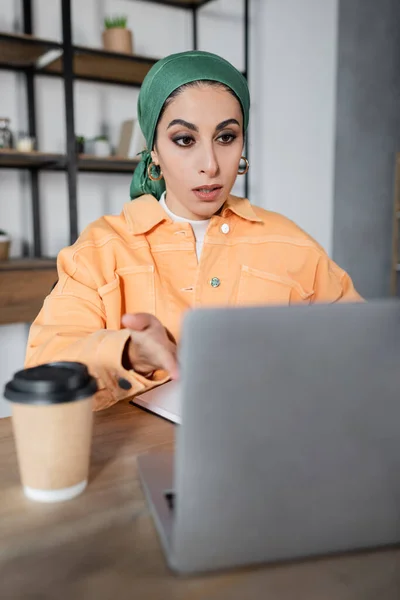 Surprised muslim woman in headkerchief pointing at blurred laptop near coffee to go — Stock Photo