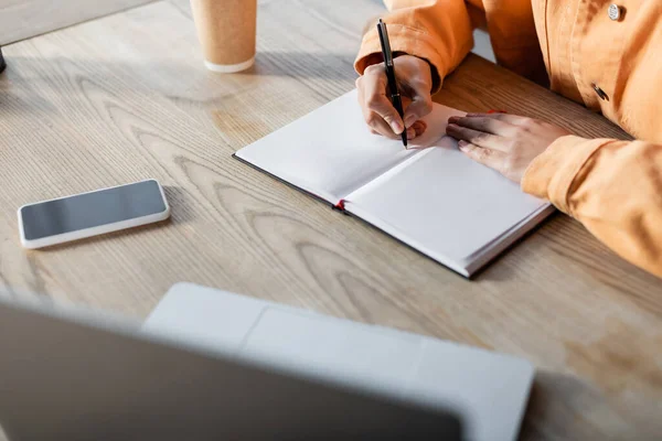 Vue partielle de l'écriture des étudiants dans un cahier vierge près d'un ordinateur portable flou et d'un smartphone sur le bureau à la maison — Photo de stock