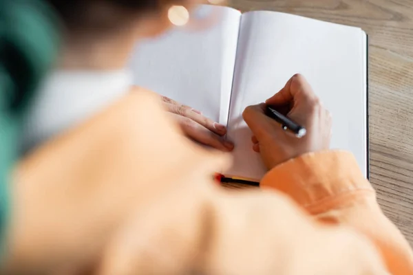 Ausgeschnittene Ansicht eines verschwommenen Schülers, der zu Hause auf leeres Notizbuch schreibt — Stockfoto
