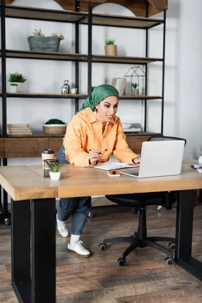 Feliz mujer musulmana sonriendo cerca de la computadora portátil, mientras que el aprendizaje en casa - foto de stock