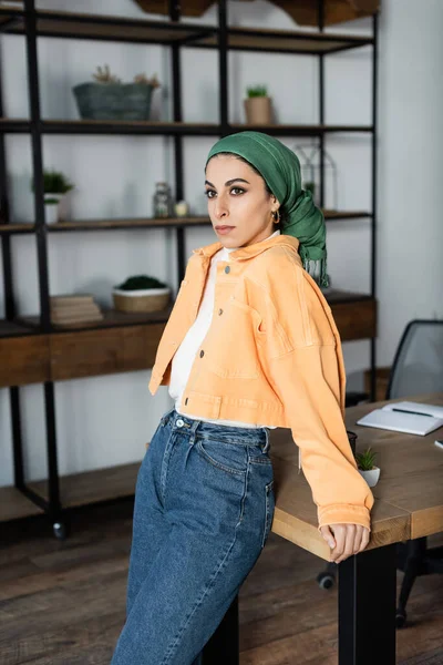 Young muslim woman in orange jacket and headkerchief standing near desk at home — Stock Photo