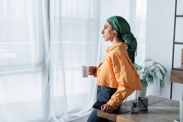 Vista laterale di donna musulmana in fazzoletto e giacca arancione in piedi con tazza di tè a casa — Foto stock