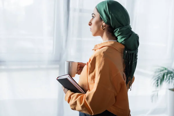 Vue latérale de l'étudiant arabe avec cahier et tasse de thé regardant loin à la maison — Photo de stock