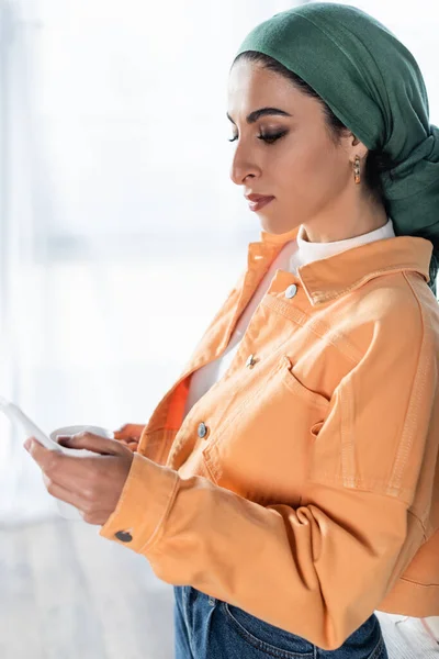 Jeune femme musulmane dans la messagerie de foulard sur smartphone à la maison — Photo de stock