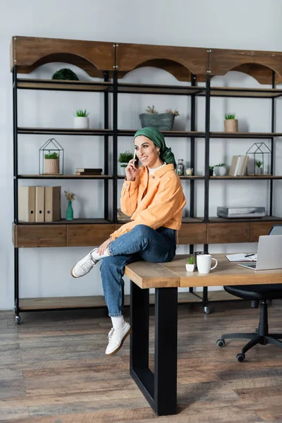 Fröhliche muslimische Frau, die auf dem Schreibtisch neben dem Laptop sitzt und mit dem Handy telefoniert — Stockfoto