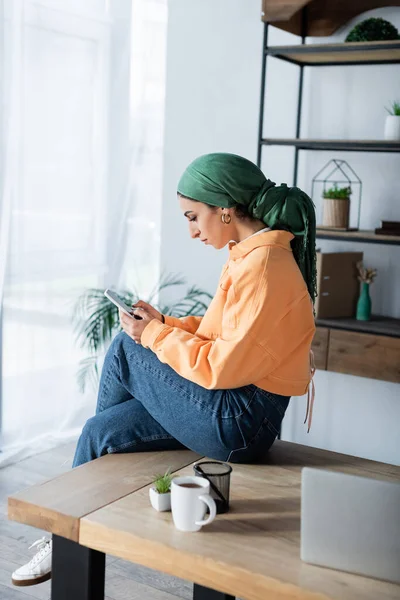 Junge muslimische Studentin in Taschentuch sitzt zu Hause am Schreibtisch und benutzt Handy — Stockfoto