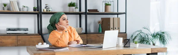 Femme musulmane regardant loin tout en étant assis près de l'ordinateur portable à la maison, bannière — Photo de stock