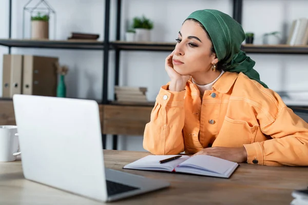 Мусульманская женщина сидит с рукой возле лица во время онлайн-урока на ноутбуке дома — стоковое фото
