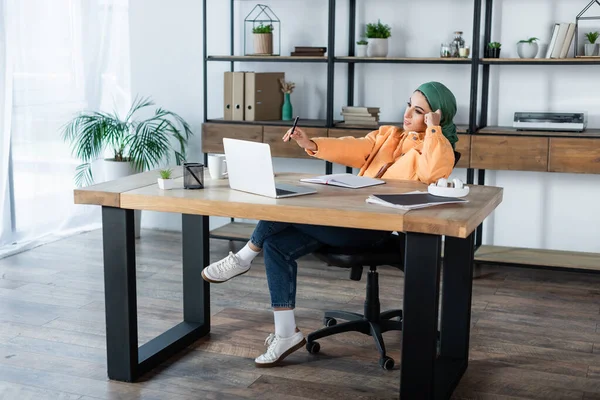 Longitud completa de la mujer musulmana sosteniendo pluma mientras se aprende cerca de la computadora portátil en la sala de estar - foto de stock