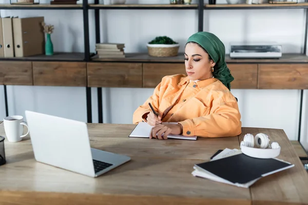 Mulher muçulmana na cabeça escrevendo em caderno enquanto estudava perto de laptop em casa — Fotografia de Stock