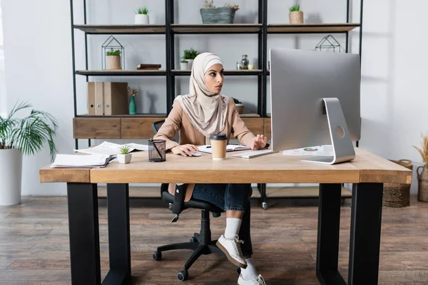 Vista completa de la mujer árabe que trabaja en la computadora cerca de la bebida para llevar y documentos en el escritorio - foto de stock