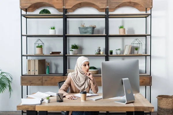 Mujer musulmana que trabaja en la computadora cerca de rack en la oficina en casa - foto de stock