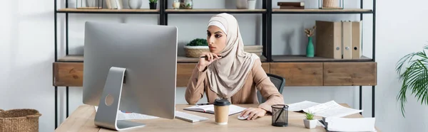 Молодая арабская женщина в хиджабе смотрит на монитор компьютера во время работы в домашнем офисе, баннер — стоковое фото