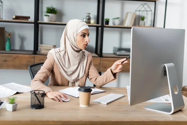 Muslim woman pointing at computer monitor while working in home office — Stock Photo