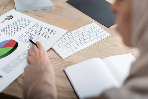 Cropped view of blurred arabian woman pointing at analytics near blank notebook — Stock Photo