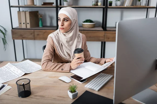 Мусульманская женщина смотрит в сторону и держит кофе, чтобы пойти, сидя в домашнем офисе — стоковое фото