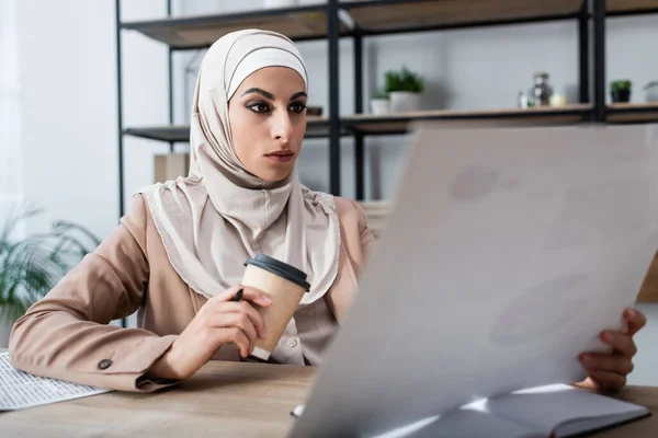 Mujer árabe en hijab sosteniendo una taza de papel y un documento borroso mientras trabajaba en casa - foto de stock