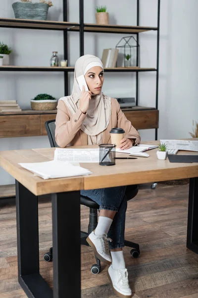 Muslimische Frau spricht auf Smartphone, während sie neben Kaffee sitzt, um zu Hause am Schreibtisch zu arbeiten — Stockfoto