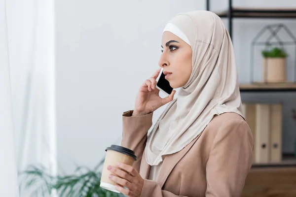 Side view of muslim woman talking on smartphone while standing with coffee to go at home — Stock Photo