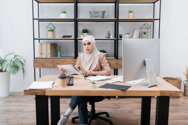 Мусульманка держит газету и смотрит в камеру возле компьютера на рабочем столе дома — стоковое фото