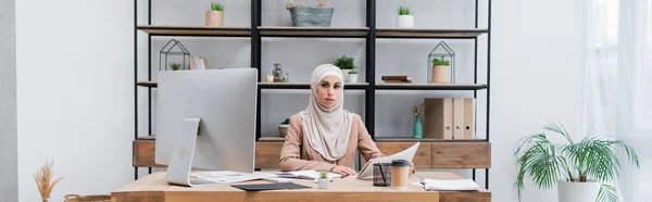 Arabian woman in hijab looking at camera near documents and computer on work desk, banner — Stock Photo