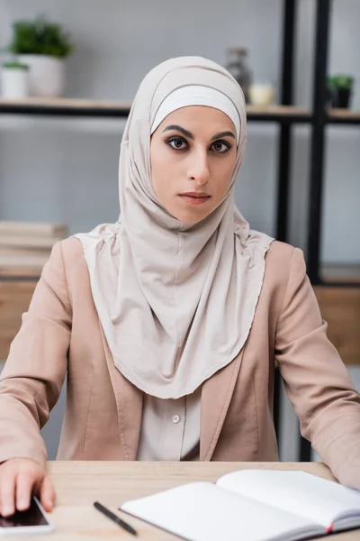 Mulher muçulmana em hijab sentado perto de notebook em branco e telefone celular em casa — Fotografia de Stock